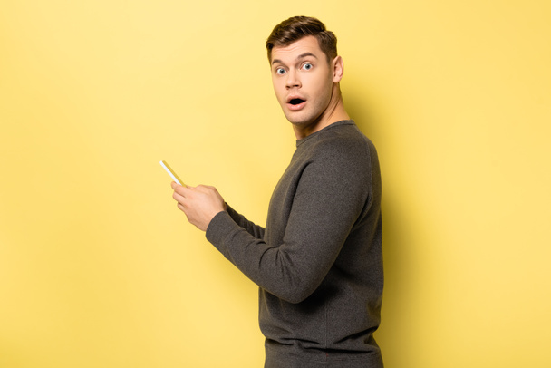 Erstaunter Mann blickt auf Kamera, während er Smartphone auf gelbem Hintergrund benutzt - Foto, Bild