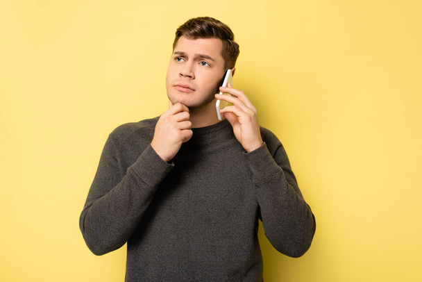Pensive man with hand near chin talking on smartphone on yellow background - Фото, изображение