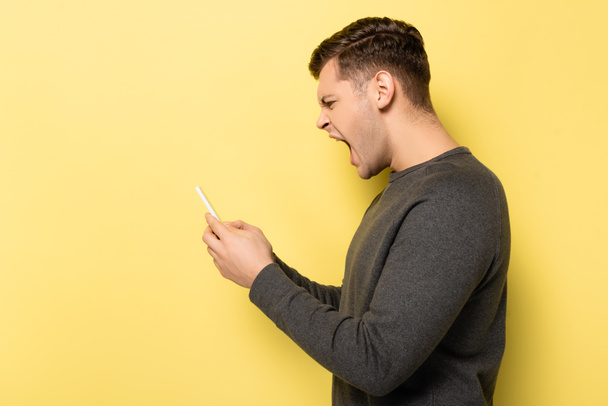Vista laterale di urla uomo utilizzando il cellulare su sfondo giallo - Foto, immagini