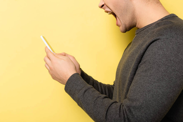 Vue recadrée de l'homme agressif en utilisant un smartphone sur fond jaune - Photo, image