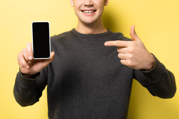 画面が空白のスマートフォンを保持し、黄色の背景に指で指している男の作物ビュー - 写真・画像