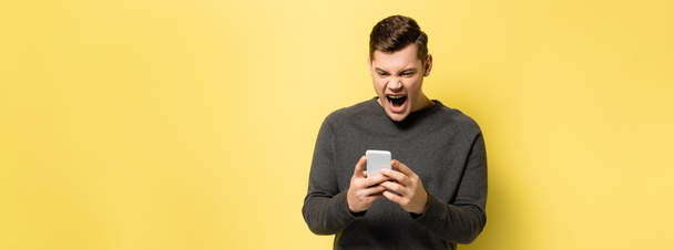 Ουρλιάζοντας και θυμωμένος άνθρωπος χρησιμοποιώντας το κινητό τηλέφωνο σε κίτρινο φόντο, πανό  - Φωτογραφία, εικόνα