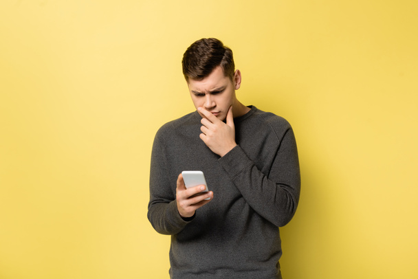 Thoughtful man looking at smartphone on yellow background - Фото, зображення