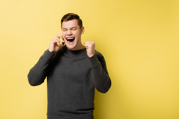 Excited man showing yes gesture while talking on smartphone on yellow background - Foto, imagen