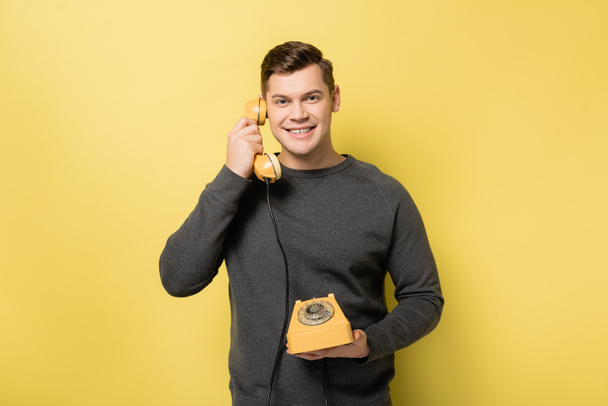 Smiling man talking on vintage telephone on yellow background - 写真・画像