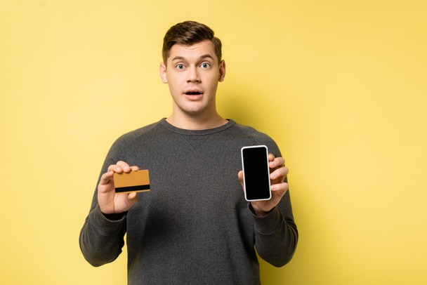 Amazed man holding credit card and smartphone on yellow background - Photo, Image