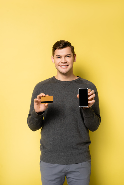 Usmívající se muž ukazující kreditní kartu a smartphone na žlutém pozadí - Fotografie, Obrázek