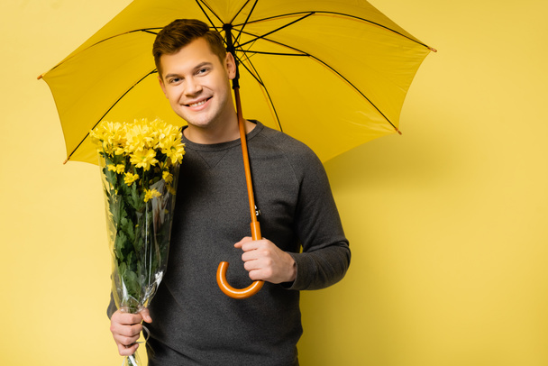 Smiling man with flowers and umbrella looking at camera on yellow background - Photo, image