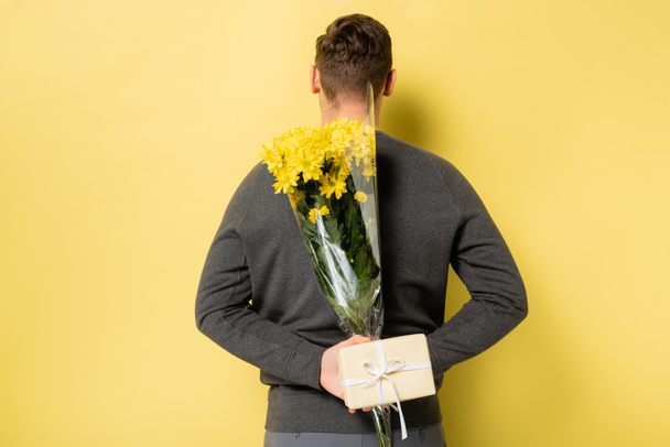 Back view of man hiding flowers and gift on yellow background - Zdjęcie, obraz