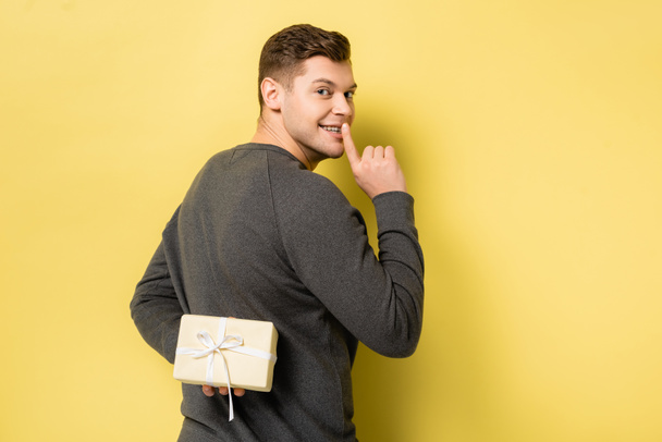 Back view of man showing shh gesture and hiding present on yellow background - Foto, imagen
