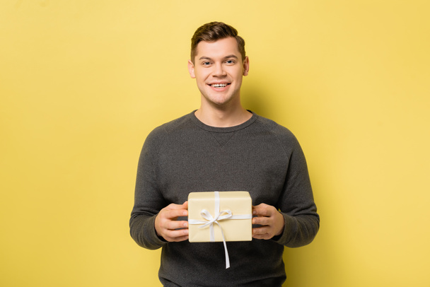 Uomo sorridente con scatola regalo guardando la fotocamera su sfondo giallo - Foto, immagini