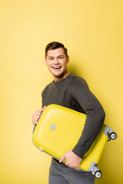 Cheerful man in grey pullover holding suitcase on yellow background - 写真・画像