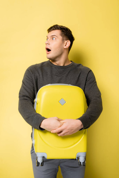 Amazed man with open mouth holding suitcase on yellow background - Фото, зображення