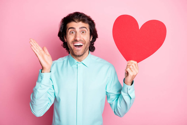 Photo portrait of shocked man holding red heart card in one hand isolated on pastel pink colored background - Foto, Bild