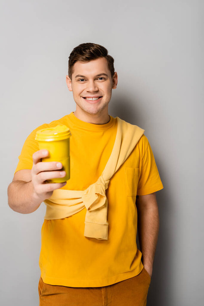 Smiling man with hand in pocket holding coffee to go on blurred foreground on grey background - Foto, imagen