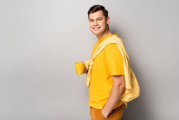 Young man smiling at camera while holding cup on grey background - Φωτογραφία, εικόνα