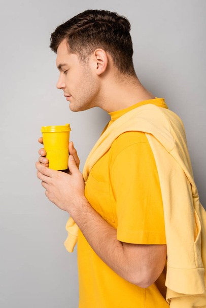 Vista lateral del hombre oliendo café para llevar sobre fondo gris - Foto, Imagen