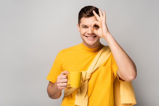 Smiling man with cup showing ok gesture on grey background - Foto, Bild