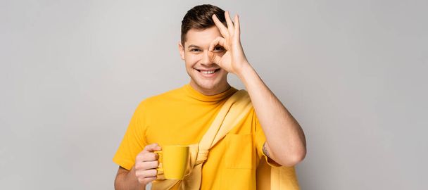 Cheerful man showing okay symbol and holding mug on grey background, banner  - Photo, Image