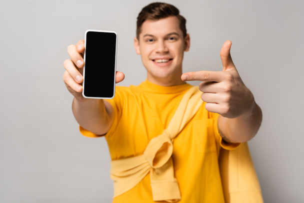 Smartphone with blank screen in hand of smiling man pointing with finger blurred on grey background - Photo, Image