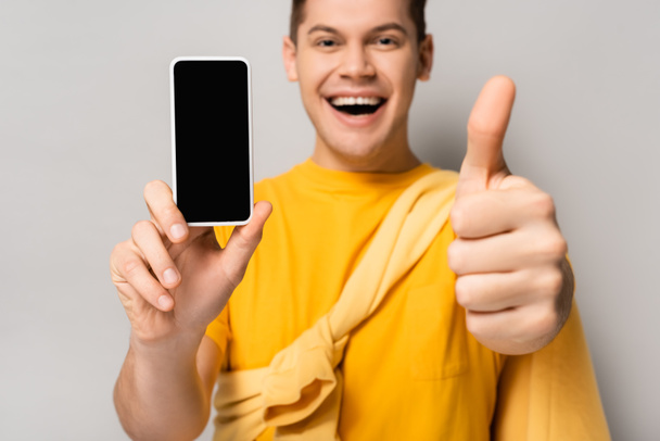 Smartfon z pustym ekranem w ręku uśmiechnięty mężczyzna pokazuje uderzenie w górę rozmyty na szarym tle - Zdjęcie, obraz