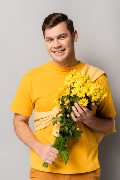 Young man smiling at camera while holding yellow chrysanthemums on grey background - Photo, image