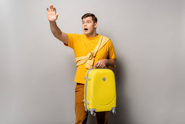 Worried traveler with suitcase waving hand on grey background - Фото, изображение