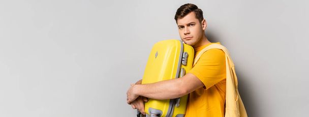 Sad traveler with suitcase looking at camera on grey background, banner  - Zdjęcie, obraz
