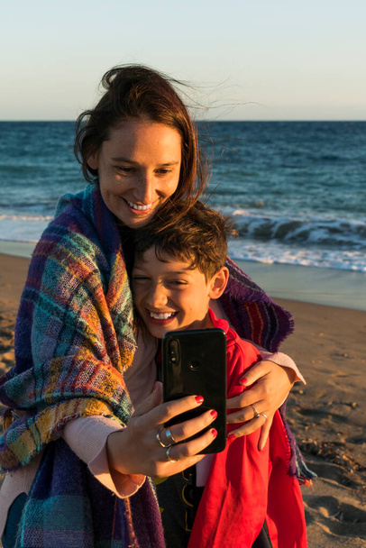 Madre e hijo están tomando fotos con el teléfono en la playa en un día ventoso - Foto, imagen