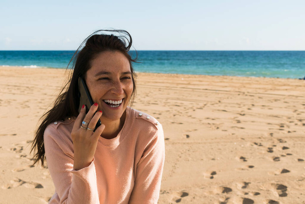 笑顔の女性は、バックグラウンドで海とビーチで彼女の携帯電話で話しています - 写真・画像
