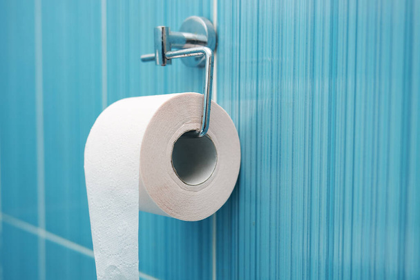 Un rotolo di carta igienica pende da un supporto metallico contro una parete di piastrelle blu.  - Foto, immagini