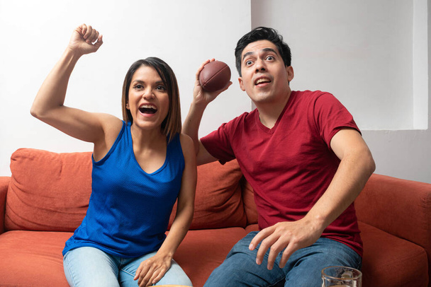 Feliz pareja latina levantando sus brazos, viendo un partido de fútbol americano. Pareja emocionada sentada en un sofá - Foto, imagen