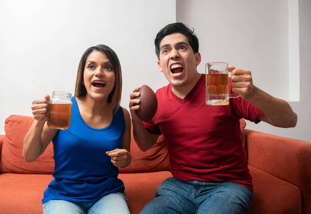 Ευτυχισμένη γυναίκα που κρατάει μια μπύρα και ενθουσιασμένος άντρας που κρατάει μια μπύρα και μια αμερικάνικη μπάλα ποδοσφαίρου. Λατινική ζευγάρι ενθουσιασμένος κατά τη διάρκεια ενός αμερικανικού ποδοσφαιρικού αγώνα - Φωτογραφία, εικόνα