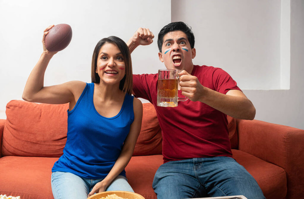 Une femme heureuse tenant un ballon de football américain et un homme excité tenant une bière. Couple latin excité lors d'un match de football américain - Photo, image