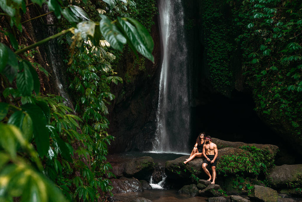 Beautiful couple at a tropical waterfall. A single couple is traveling on the island of Bali in Indonesia. A woman and a man at a beautiful waterfall. Vacation on a tropical island. Copy space - Valokuva, kuva
