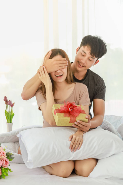 Concetto di coppia e San Valentino. Sorridente uomo asiatico sorpresa la sua ragazza con bella scatola regalo sul letto in camera da letto. - Foto, immagini