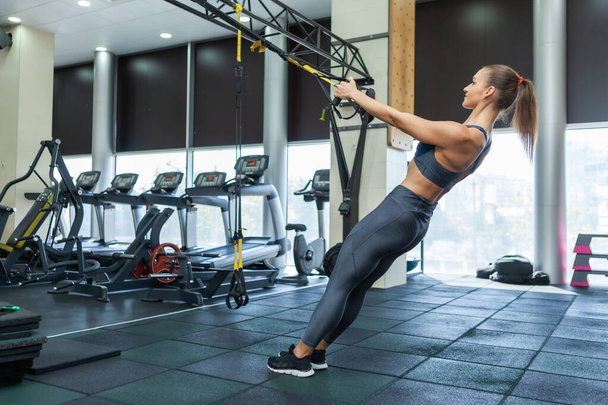 Jonge aantrekkelijke gespierde vrouw in sportkleding traint spieren met trx weerstand systeem in een moderne fitnessruimte. Kruistraining. Moderne fitness - Foto, afbeelding