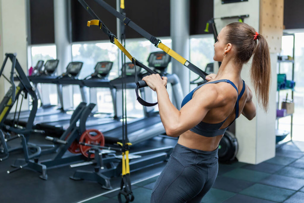 Young attractive muscular woman in sportswear trains muscles with trx resistance system in a modern gym. Cross training. Modern fitness - Photo, image