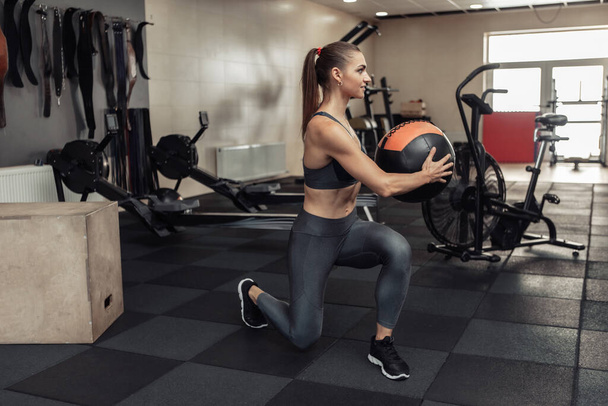 Mujer joven en forma de cruz haciendo ejercicio con balón de medicina en el gimnasio moderno. Entrenamiento funcional. Estilo de vida saludable - Foto, Imagen