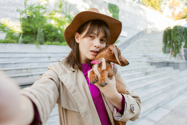 Young attractive woman with dachshund puppy in city park makes selfie portrait - Photo, Image