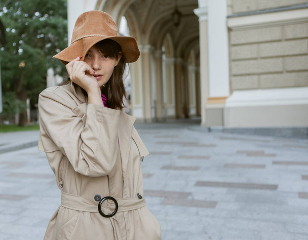Modeporträt einer jungen Frau mit Filzhut und herbstlichem Trenchcoat in der Stadt - Foto, Bild