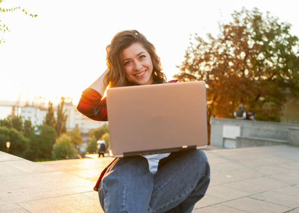 Молодая веселая студентка, сидящая с ноутбуком на улице рано утром на рассвете - Фото, изображение