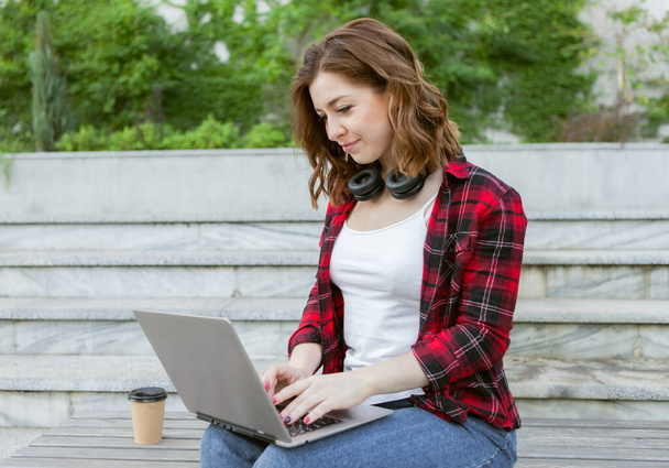 若い陽気な女性学生は屋外ノートパソコンを使用しています。遠隔作業又は教育の概念 - 写真・画像