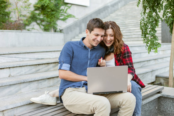 Young cheerful couple of lovers spend time together while use laptop outdoors. Happy, funny people. Love concept. - Zdjęcie, obraz