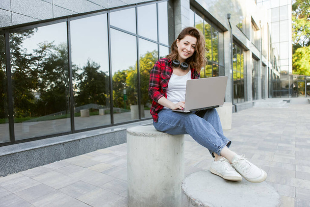 Young cheerful woman student using laptop outdoors. Distance work or education concept - Photo, Image