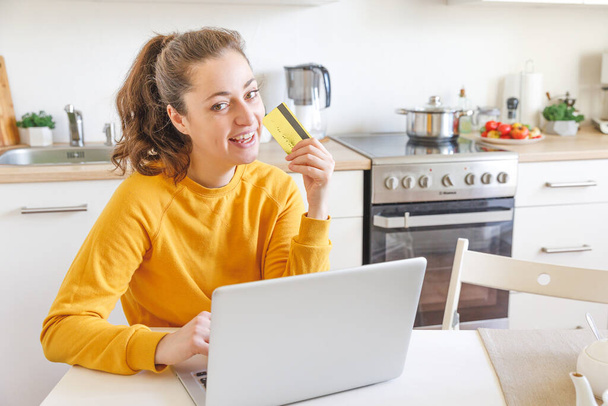 Woman shopping online and paying with gold credit card. Young girl sitting with laptop buying on Internet enter credit card details on kitchen indoor background. Online shopping e-commerce concept - Photo, Image