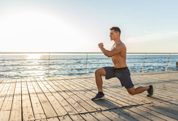 Hombre musculoso y guapo con el torso desnudo haciendo ejercicios de embestida en la playa al amanecer. Estilo de vida saludable - Foto, imagen