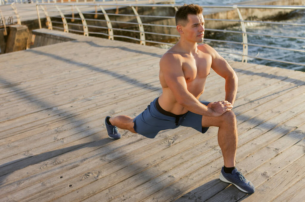 Giovane uomo muscoloso con torso nudo facendo gamba che si estende sulla spiaggia all'alba - Foto, immagini
