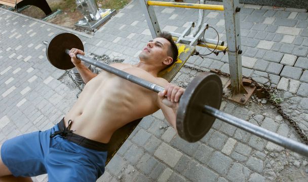 Muscular man doing barbell bench press on a bench outdoors. Healthy lifestyle. Outdoor training - Photo, Image