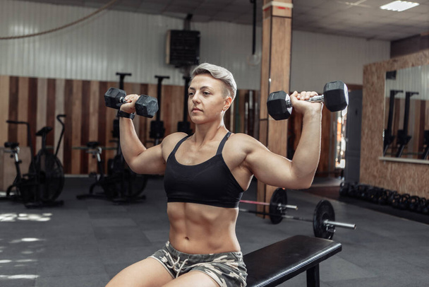 Young athletic strong woman trains the muscles of the shoulder with dumbbells in her hands while sitting on a bench in a cross gym. Bodybuilding and Fitness - Foto, imagen